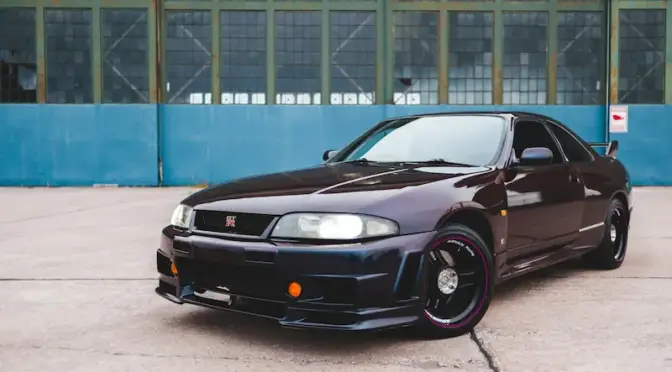 Ciemnofioletowy Nissan Skyline R33 po tuningu wizualnym, zaparkowany przed industrialnym budynkiem.